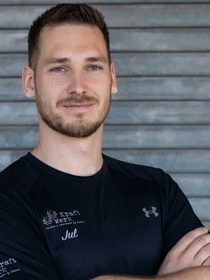 Ein Mann mit kurzem Haar lächelt leicht vor einer grauen Wand in einem schwarzen T-Shirt.